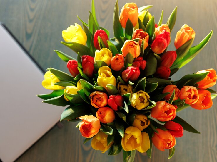 Bunter Tulpenstrauß in Gelb, Orange und Rot liegt auf Holztisch neben geschlossenem silberfarbenem Laptop im Sonnenlicht.