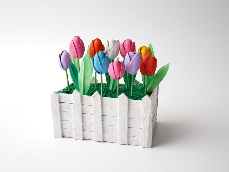 Bunt bemalte Tulpen aus Papier stehen in grünem Kunstgras, eingefasst von einem weißen Miniaturzaun aus Holz.