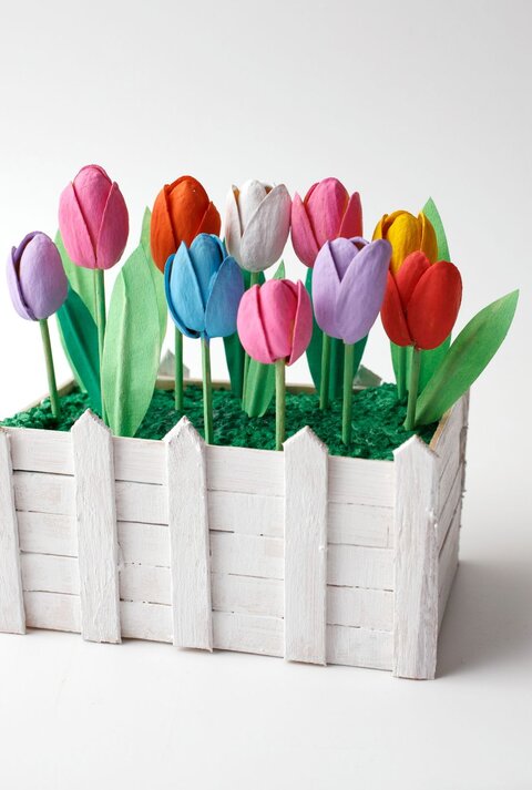 Bunt bemalte Tulpen aus Papier stehen in grünem Kunstgras, eingefasst von einem weißen Miniaturzaun aus Holz.