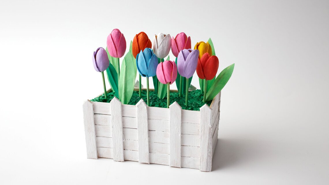 Bunt bemalte Tulpen aus Papier stehen in grünem Kunstgras, eingefasst von einem weißen Miniaturzaun aus Holz.