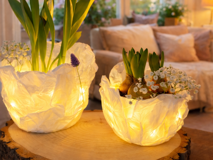 Zwei leuchtende Tulpen aus Butterbrotpapier stehen mit Frühlingsblumen auf einer Baumscheibe in gemütlich beleuchtetem Wohnzimmer.