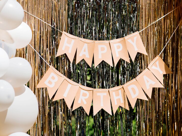 Geburtstagsgirlande basteln: Beige Wimpelkette mit weißem „Happy Birthday“-Schriftzug hängt festlich vor schimmerndem Goldvorhang mit Ballondekoration.