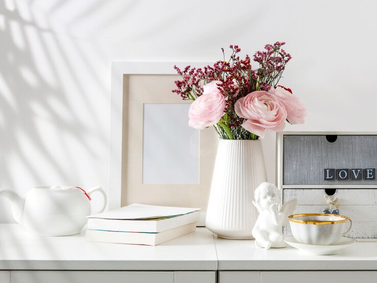 Zartrosa Ranunkeln und pinke Schleierkrautblüten in weißer Vase auf Kommode mit Teetasse, Büchern und Dekoengel.