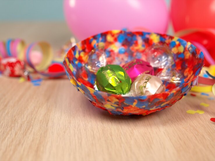 Farbenfrohe Konfetti-Schale mit Süßigkeiten auf hellem Tisch, daneben Schere, Papierstreifen und Luftballons in fröhlicher Partyatmosphäre.