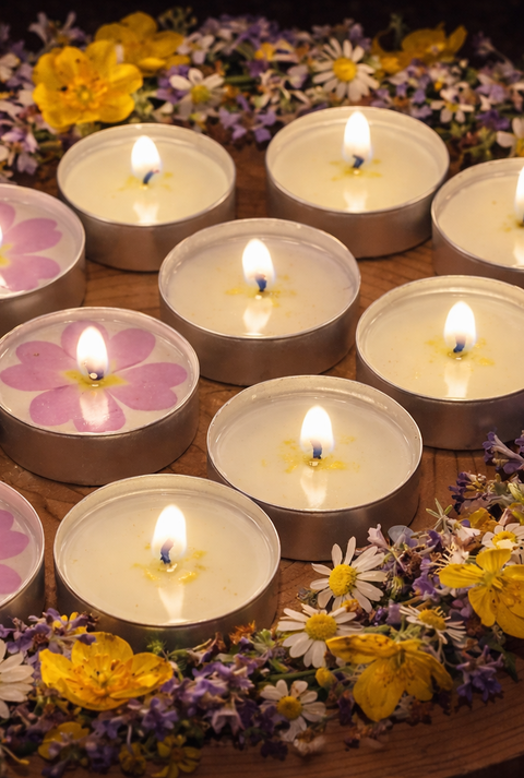 Mehrere brennende Teelichter mit rosa Primeln liegen kreisförmig auf Holz, umrahmt von bunten Wiesenblumen.