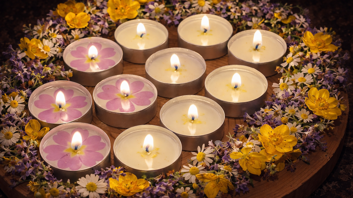 Mehrere brennende Teelichter mit rosa Primeln liegen kreisförmig auf Holz, umrahmt von bunten Wiesenblumen.