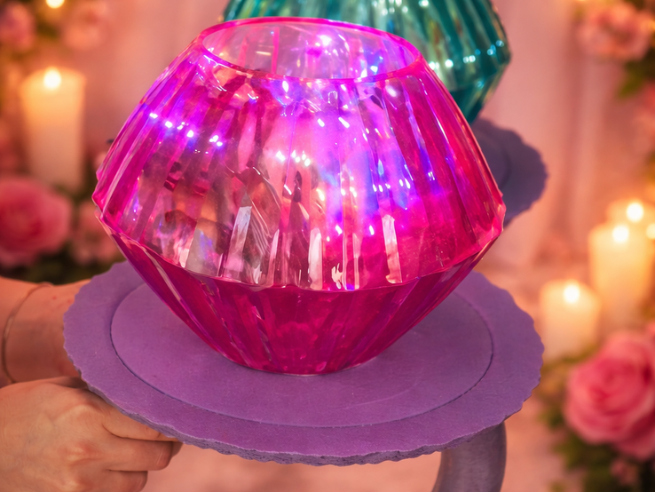 Ein selbst gebastelter Diamantring aus pinkem und türkisfarbenem Glas leuchtet auf einer lilafarbenen Halterung vor Kerzen und Rosen.