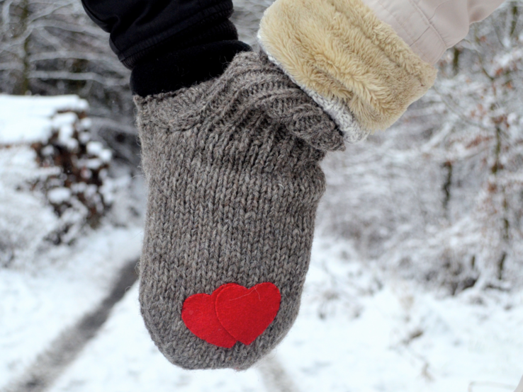 Zwei Hände in grauem Doppelhandschuh mit roten Herzen halten sich auf einem verschneiten Waldweg im Winter.