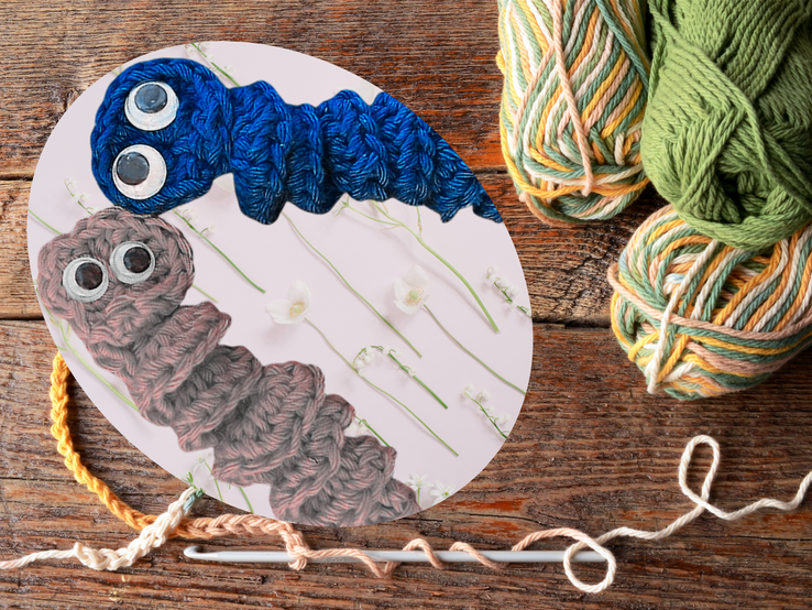 Zwei gehäkelte Würmchen mit großen Augen aus Wolle in Blau und Braun liegen auf pastellfarbenem Hintergrund mit weißen Blumen.