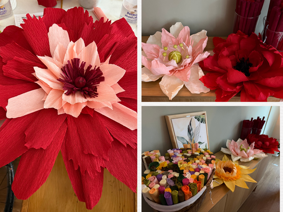 Große rote und rosafarbene Krepppapierblumen liegen neben bunten Papierrollen und gerahmter Pflanzenillustration auf Holzkommode. | © Madeleine Liedke 