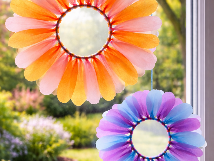 Zwei blütenförmige Blumenkränze hängen als Fensterdekoration in Orange und Violett  vor einem blühenden Frühlingsgarten mit Tulpen und Weg.