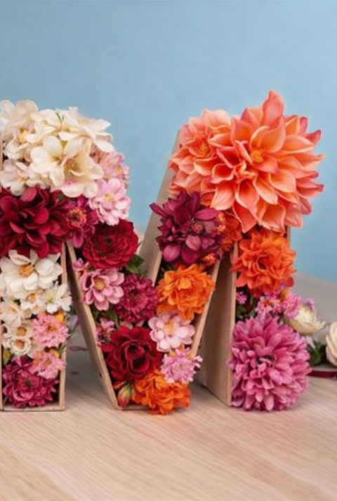 Mit bunten Dahlien und Rosen gefüllter Buchstabe M aus Karton steht auf Holztisch vor hellblauem Hintergrund.