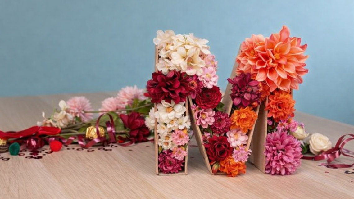 Mit bunten Dahlien und Rosen gefüllter Buchstabe M aus Karton steht auf Holztisch vor hellblauem Hintergrund.