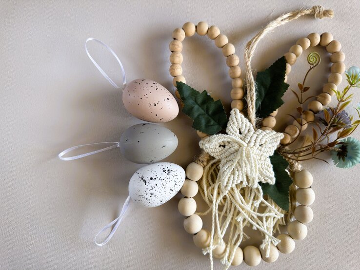 Drei pastellfarbene Ostereier mit weißen Bändern liegen neben einem Holzperlenherz mit Makramee-Stern und grünen Blättern.