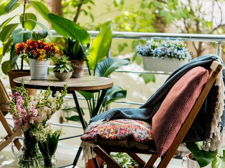 Ein gemütlicher Balkon mit Holztisch, bunten Blumentöpfen, gemustertem Kissen und Decke auf Klappstuhl vor üppigem Grün.