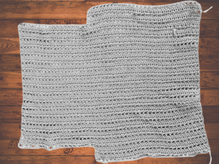 Rechteckiges, weißes Häkelstück aus feinem Garn, locker gehäkelt, auf einem Tisch aus Holz mit warmer Holzmaserung platziert. | © Canva [M]