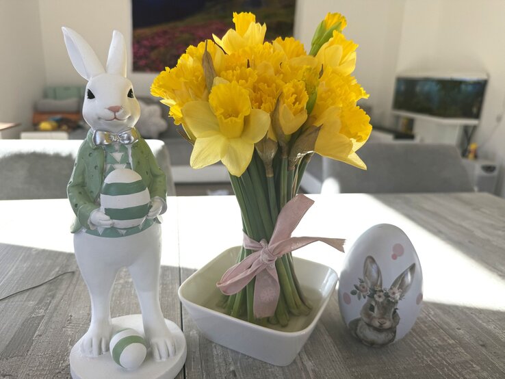 Gelbe Narzissen in kleiner Vase mit rosa Schleife stehen auf Holztisch neben Osterhasenfigur mit Ei und bemaltem Deko-Ei im hellen Wohnzimmer.