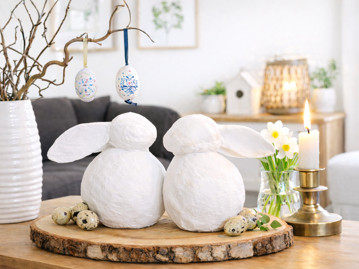 Zwei weiße DIY-Deko-Osterhasen aus Zeitung sitzen Rücken an Rücken auf einer Holzscheibe, daneben Kerze, Narzissen und kleine Wachteleier.