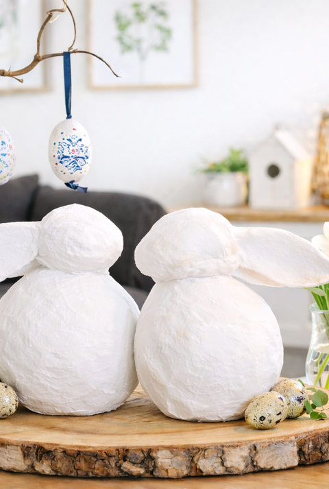 Zwei weiße DIY-Deko-Osterhasen aus Zeitung sitzen Rücken an Rücken auf einer Holzscheibe, daneben Kerze, Narzissen und kleine Wachteleier.