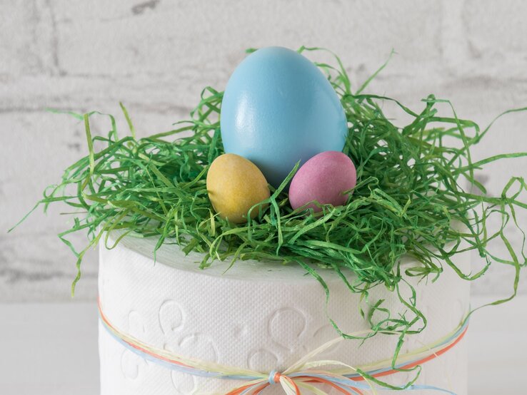 Ein großes blaues Osterei und zwei kleine gelbe und rosafarbene Eier liegen in grünem Ostergras auf einer dekorierten Toilettenpapierrolle.