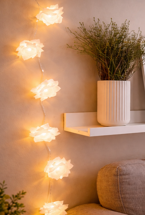 Warme Lichterkette aus Butterbrotpapier mit weißen Blüten hängt neben Wandregal mit Vase, Bild und Pflanze über einem beigen Sofa im gemütlichen Wohnzimmer.