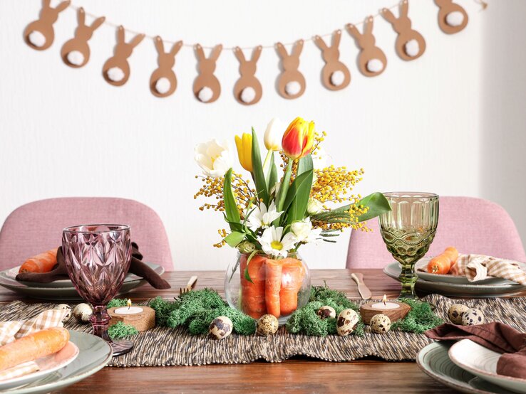 Frühlingshaft gedeckter Tisch mit Tulpenstrauß in Karottenvase, bunten Gläsern, Möhren auf Tellern und Hasengirlande im Hintergrund.