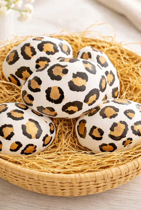 Dekorierte Ostereier im Leopardenlook ruhen in einem runden Korb aus Naturmaterial, umgeben von feinem Stroh auf heller Holzfläche.