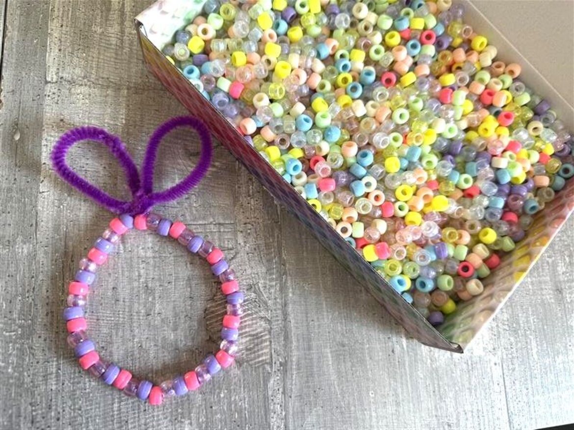 Runder Perlenring in Rosa und Lila mit violetter Pfeifenputzerschleife in Osterhaseform liegt neben offener Box voller bunter Bastelperlen nah. | © Madeleine Liedke 