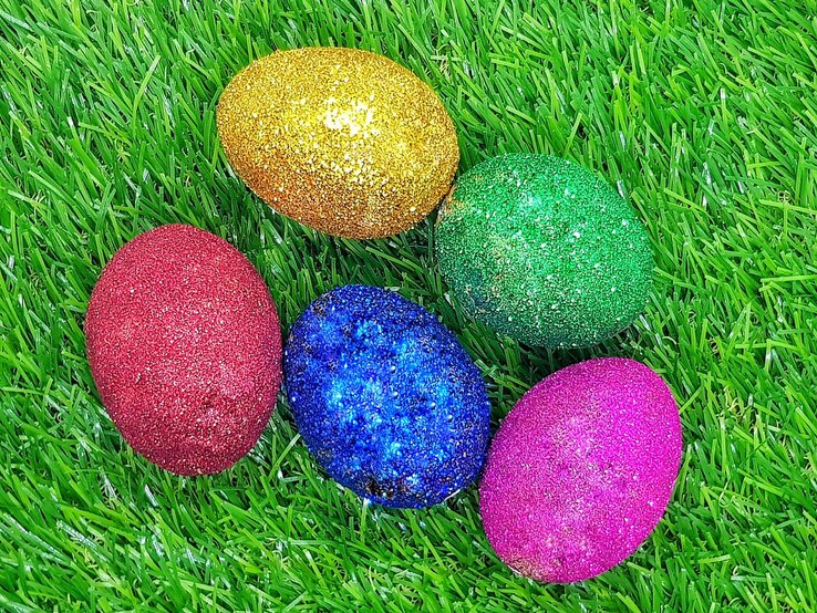 Fünf glitzernde Ostereier in Rot, Blau, Grün, Gold und Pink liegen dicht beieinander auf frischem, leuchtend grünem Gras.