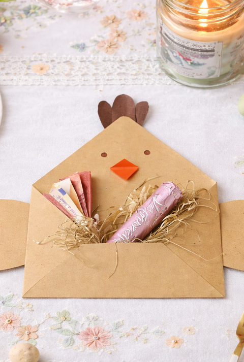 Gebastelter Hennen-Briefmschlag als Geldgeschenk mit Geldscheinen und Schokoriegel auf hellen Tisch umgeben von Tulpen, Kerze und bunten Ostereiern.