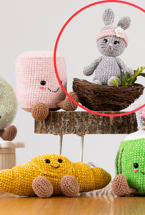 Ein gehäkelter grauer Hase mit rosa Stirnband und Blüte sitzt in einem kleinen Vogelnest, umgeben von grünen Pflanzen auf einem Holzpodest.