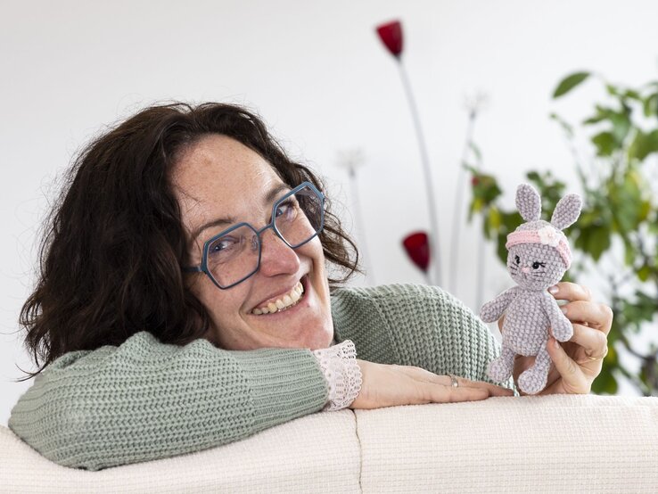 Eine Frau in grünem Strickpulli hält einen kleinen, gehäkelten grauen Hasen mit rosa Stirnband und Blüte vor hellem Hintergrund. | © BILD der FRAU/Photoproduction Bernd Hanselmann