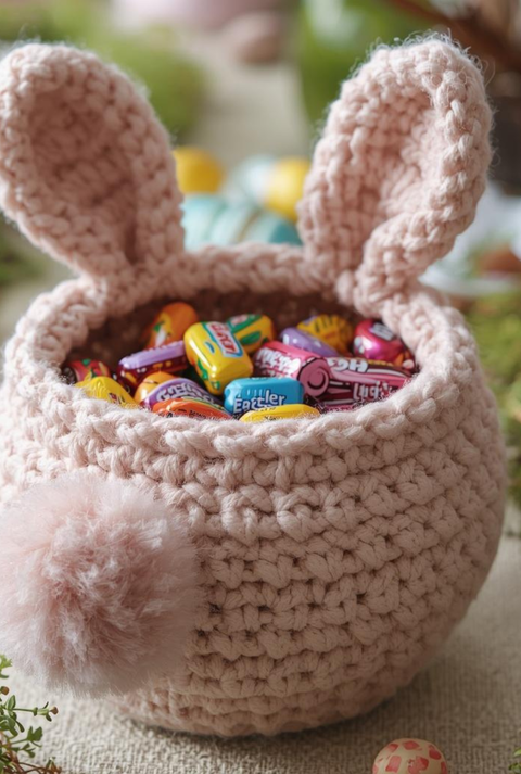 Ein gehäkeltes Körbchen mit Hasenohren, gefüllt mit bunter Oster-Süßigkeiten, steht von Eiern und Frühlingsgrün umgeben auf einem Tisch.