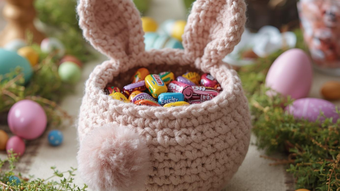 Ein gehäkeltes Körbchen mit Hasenohren, gefüllt mit bunter Oster-Süßigkeiten, steht von Eiern und Frühlingsgrün umgeben auf einem Tisch.