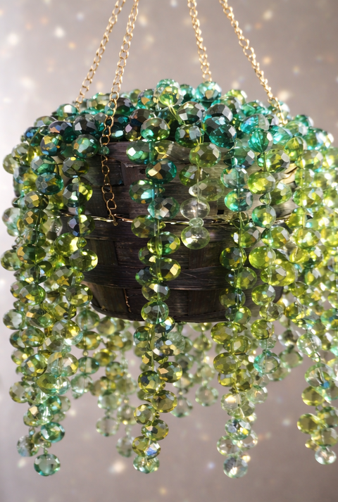 Hängender Topf an goldenen Ketten, dicht behängt mit grünen Glasperlen, die wie eine selbstgebastelte Zimmerpflanze wirken.
