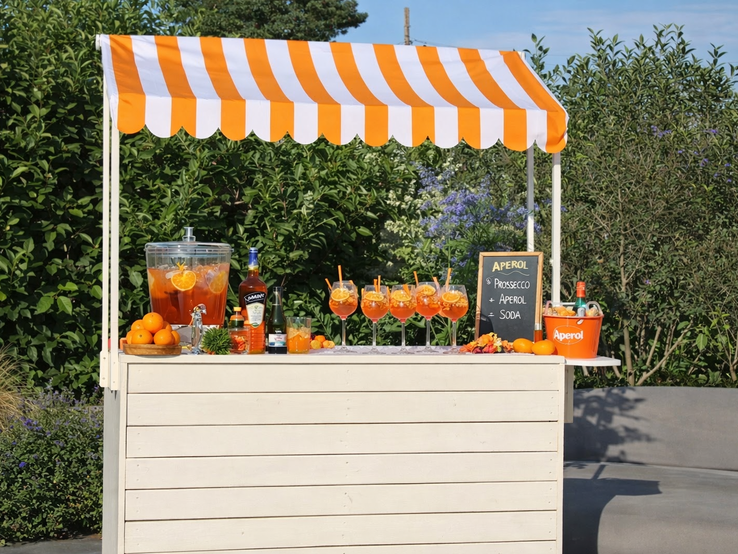 Ein mobiler Cocktailstand unter orangeweißer Markise zeigt gefüllte Aperolgläser, Spender mit Zitrusfrüchten und Deko Garten.
