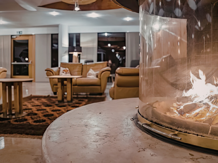 Moderner Loungebereich mit rundem Tischkamin aus Glas im Vordergrund, dahinter Ledersessel, Teppich und warme Beleuchtung.