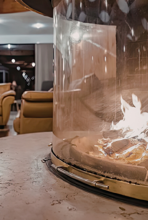 Moderner Loungebereich mit rundem Tischkamin aus Glas im Vordergrund, dahinter Ledersessel, Teppich und warme Beleuchtung.