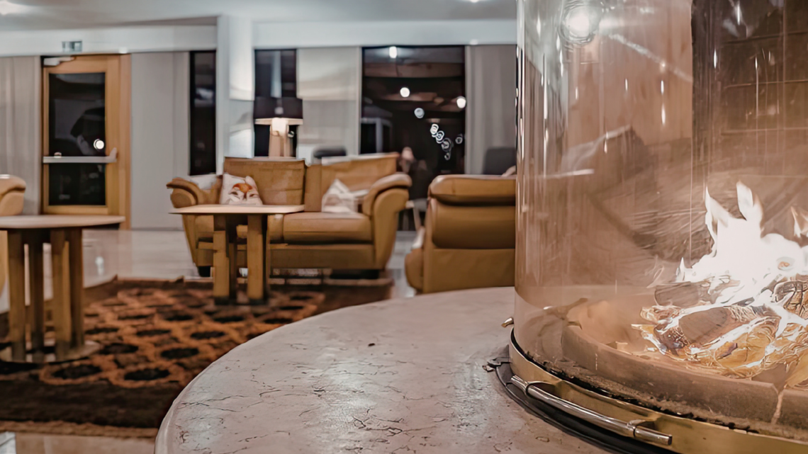 Moderner Loungebereich mit rundem Tischkamin aus Glas im Vordergrund, dahinter Ledersessel, Teppich und warme Beleuchtung.