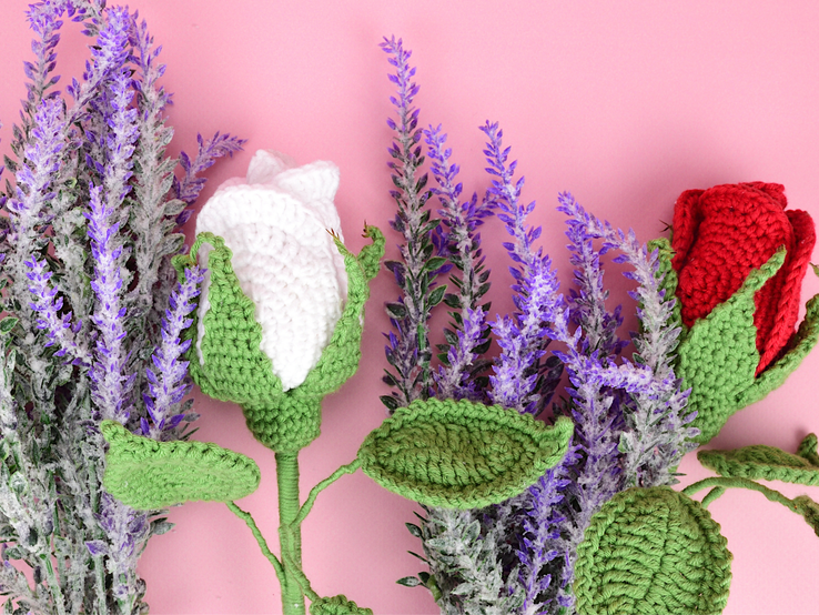 Handgehäkelte Blume mit grünen Blättern, weißen und roten Blüten vor einem Arrangement aus lilafarbenem Lavendel auf rosa Hintergrund.