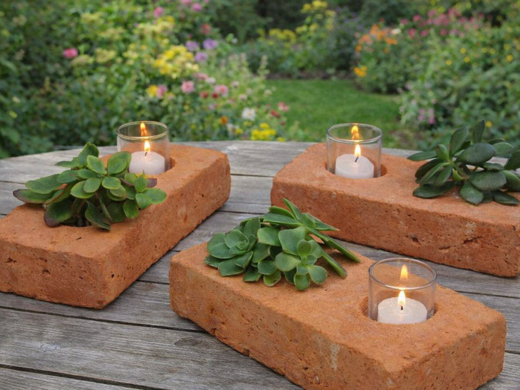 Ziegelsteine als Kerzenhalter mit Teelichtern und Sukkulenten ruhig auf Holztisch, vor üppigem Garten mit bunten Blumen.