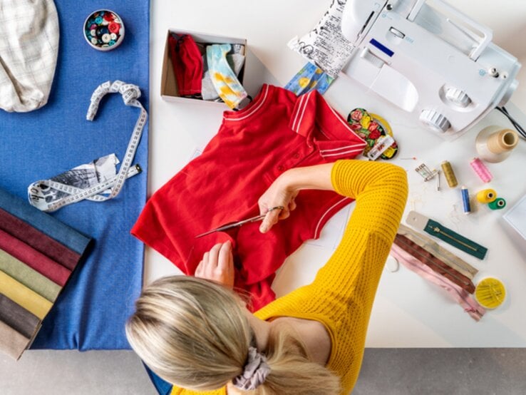 Frau schneidet altes rotes T-Shirt mit Schere um. Auf dem Tisch liegen bunte Stoffproben, Nähgarn, Knöpfe und eine Nähmaschine.