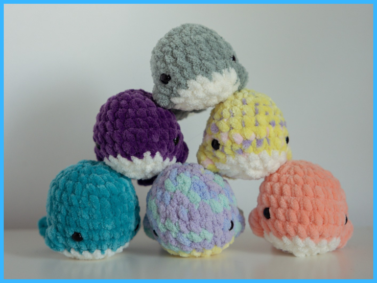 Sechs kleine, bunte, gehäkelte Stoffwale aus flauschiger Wolle bilden eine Pyramide auf weißem Untergrund vor neutraler Wand.