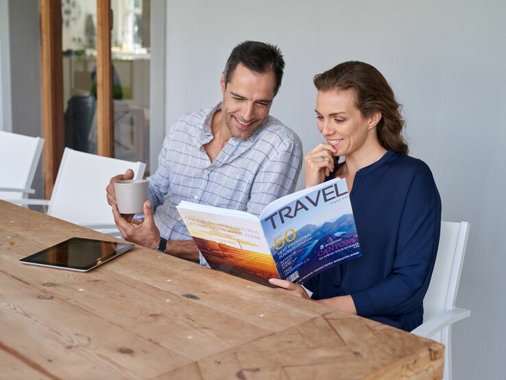 Ein Mann, der ein blau-kariertes Hemd trägt, und eine Frau, die eine dunkelblaue Bluse trägt, sitzten an einem Holztisch und schauen sich ein Reisemagazin an.