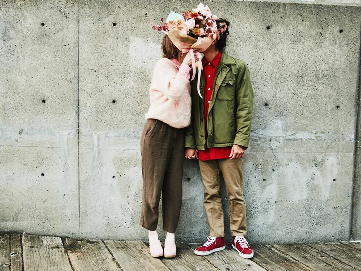 Das Bild zeigt ein Paar, das sich in einer liebevollen Pose gegenübersteht. Die Person auf der linken Seite trägt eine rosa Strickjacke, eine braune Hose und weiße Schuhe. Diese Person hält einen großen, bunten Blumenstrauß, der ihr Gesicht verdeckt. Die Person auf der rechten Seite trägt eine olivgrüne Jacke, ein rotes Hemd, braune Hosen und rote Turnschuhe. Auch das Gesicht dieser Person ist durch den Blumenstrauß verdeckt, was eine spielerische und romantische Atmosphäre schafft. Sie stehen vor einer Betonwand und auf einem hölzernen Untergrund, was einen interessanten Kontrast zur Wärme und Farbigkeit des Blumenstraußes bildet. Das Bild könnte symbolisch für die Anonymität und Universalität der Liebe stehen, oder es könnte einfach ein kreativer Ausdruck von Zuneigung sein.