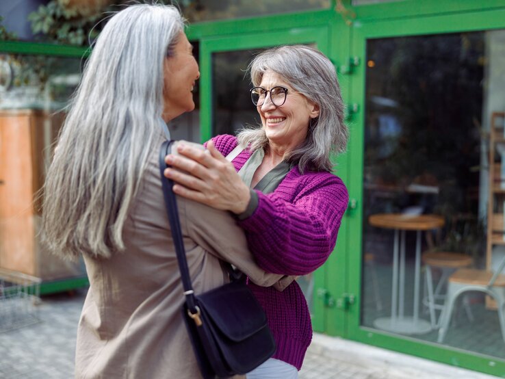 Zwei ältere Frauen mit grauen Haaren umarmen sich herzlich auf einer Straße. Die eine Frau trägt eine lila Strickjacke und Brille und lacht, während sie die andere Frau ansieht, die ihr den Rücken zuwendet. Im Hintergrund ist der Eingang zu einem Gebäude mit grünen Türen und Fenstern zu sehen, was auf ein gemütliches Café oder Geschäft hinweist