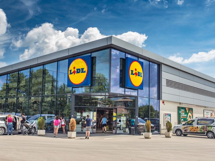 Das Foto zeigt den Eingangsbereich einer Lidl-Filiale an einem sonnigen Tag. Zwei große Schilder mit dem Lidl-Logo sind deutlich an der Fassade angebracht, was die Markenidentität des Discounters unterstreicht. Vor dem Geschäft sieht man einige Kunden, die das Geschäft betreten oder verlassen, was auf einen gewöhnlichen und belebten Einkaufstag hindeutet. Die geparkten Autos und das helle Wetter tragen zu der alltäglichen Szene eines Supermarktes bei, der für seine wirtschaftlichen Preise und ein vielfältiges Angebot bekannt ist.