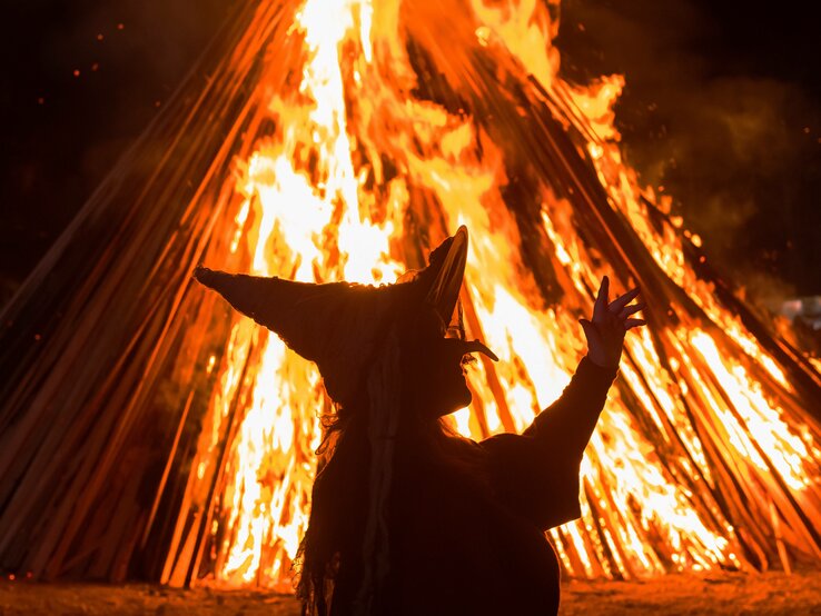 Zu sehen ist das Konterfei einer Hexe, die in der Nacht vor einem hellen, orangenen Feuer tanzt