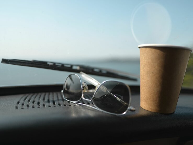 Sonnenbrille und brauner Pappbecher auf Armaturenbrett eines Autos mit Blick auf eine unscharfe Landschaft.