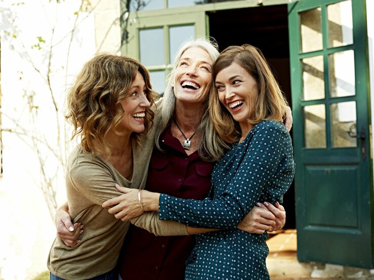 Drei lachende Frauen umarmen sich freudig vor einem offenen Fenster: eine junge Blonde, eine ältere Grauhaarige und eine Brünette